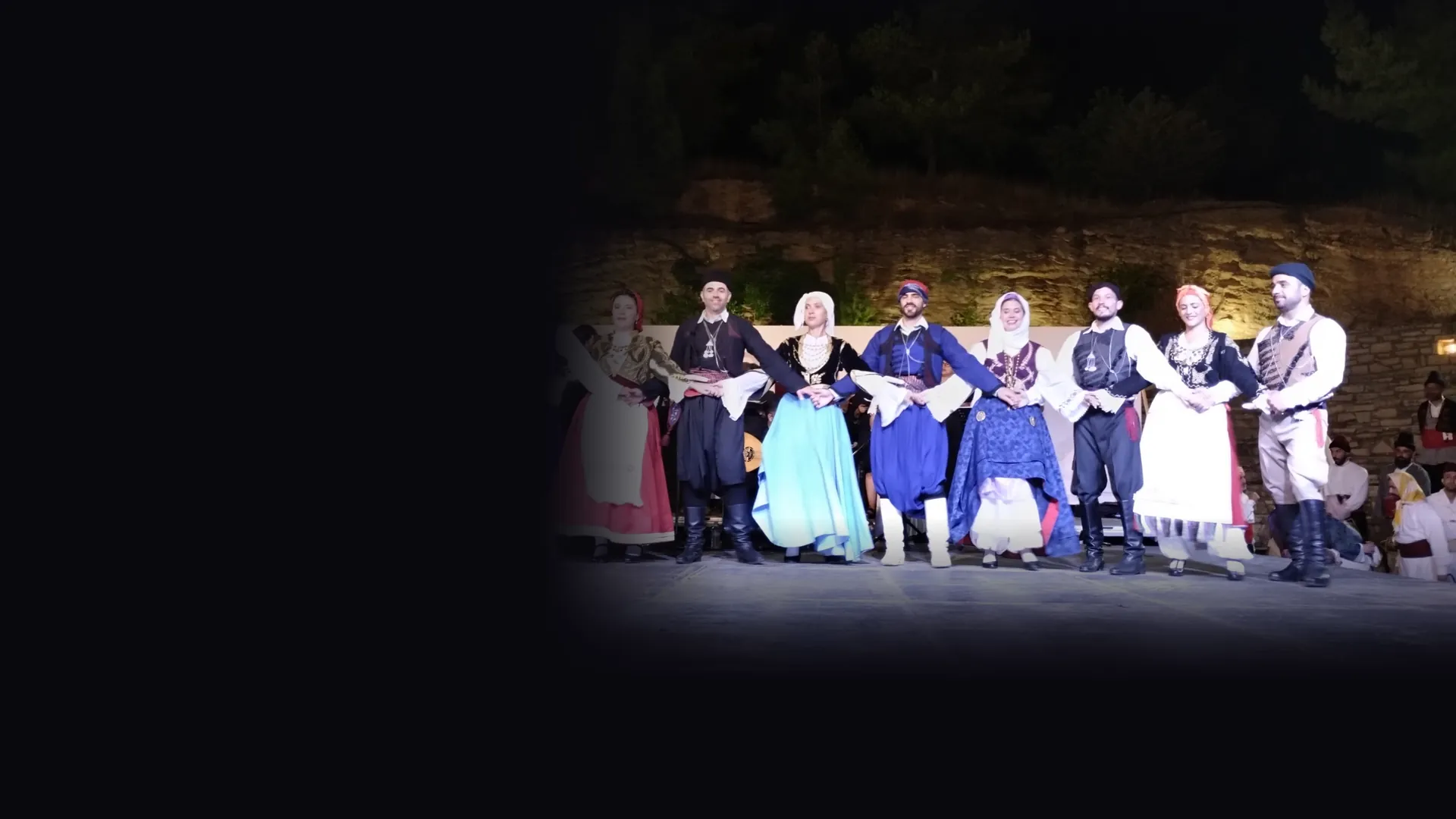 Dancers in Cretan costume stand in a line with linked hands on an open-air stage at night, in front of a lit stone terrace and pine trees