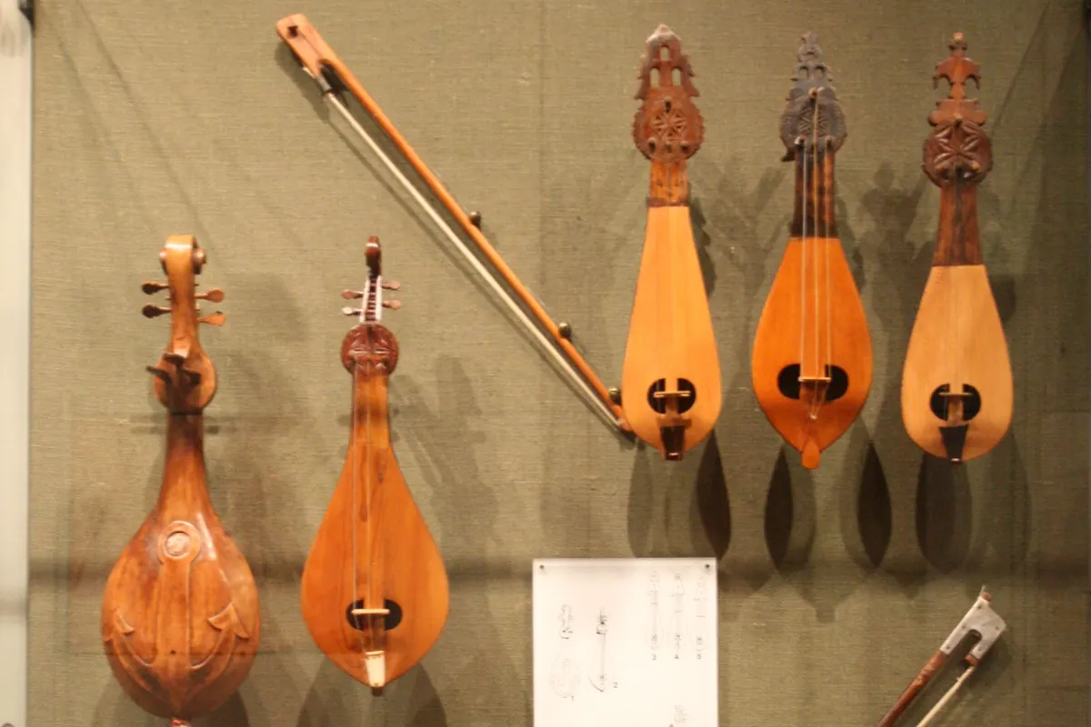 Several pear-shaped Cretan lyras with carved heads hanging on a green display wall, next to a bow